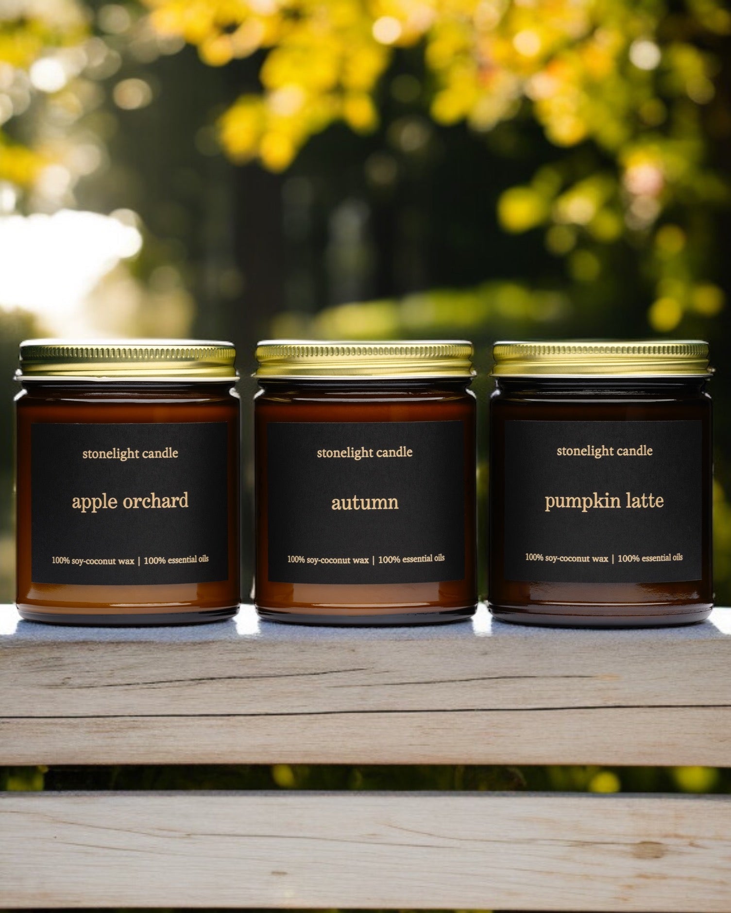 Three candles labeled 'apple orchard', 'autumn', and 'pumpkin latte' on a wooden surface with a blurred natural background.